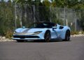 A light blue sports car with a gray stripe, reminiscent of the million-dollar cars at the Mecum Monterey 2025 Auction, is parked on a paved road near a chain-link fence and greenery.