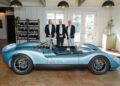Three men in suits stand behind a light blue vintage-style sports car parked indoors near shelves of wine bottles.