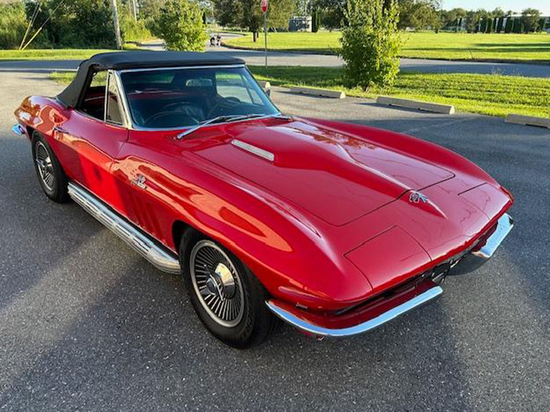 A red classic convertible sports car with a black soft top, parked on a paved surface near grass and trees, stands ready for the Fall Carlisle Collector Car Auction.