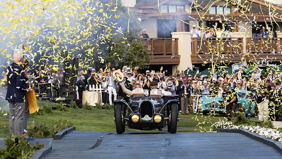 A vintage car drives onto the stage at Monterey Car Week as yellow and white confetti falls, with a crowd of people cheering in the background—a thrilling moment presented by duPont REGISTRY in 2025.