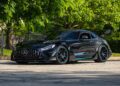 A black Mercedes-AMG sports car with a rear wing, one of the million-dollar cars expected at the Monterey 2025 Auction by Mecum, is parked on a sunlit pavement near a fence and green trees.