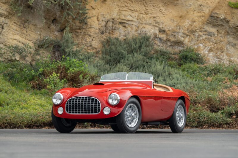 A classic red convertible sports car, perfect for Monterey Car Week 2025, is parked on a paved road with greenery and a rocky hillside in the background.