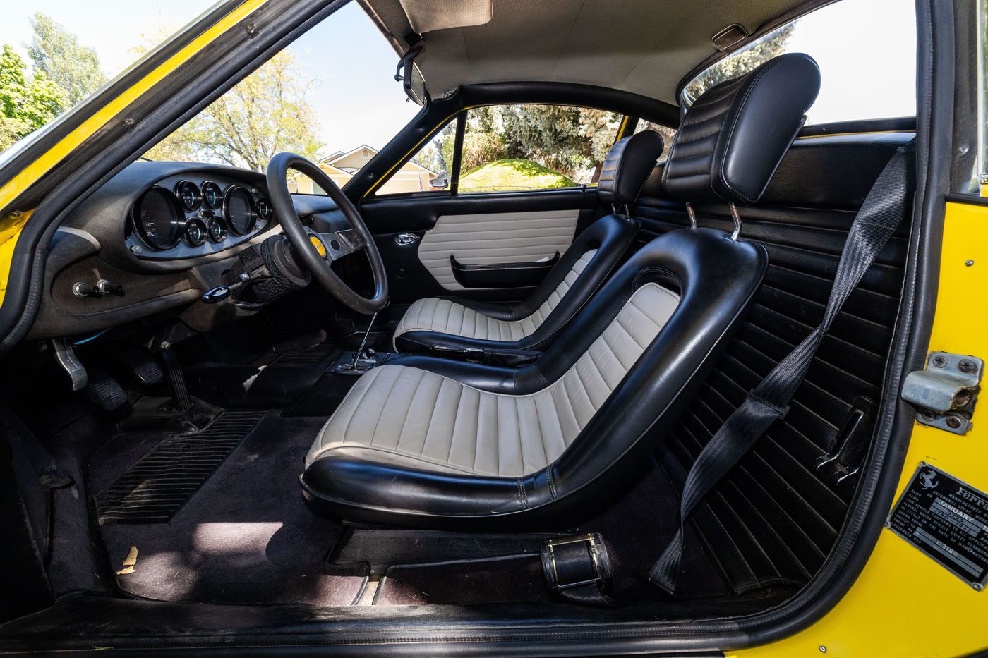 The interior of a classic car with black and white bucket seats, a black dashboard with round gauges, and a yellow exterior visible on the door frame—a true Ferrari Legends style destined for Mecum’s 2025 Auction in Monterey.