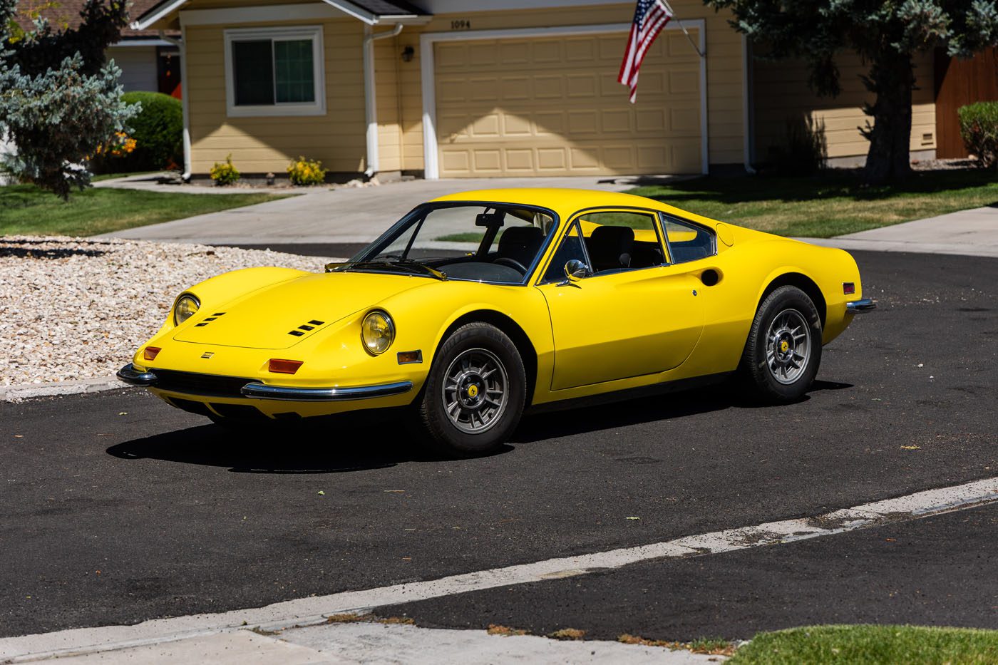A classic yellow Ferrari Legends sports car is parked on a residential street in front of a beige house with a two-car garage and an American flag, capturing the spirit of Monterey 2025.