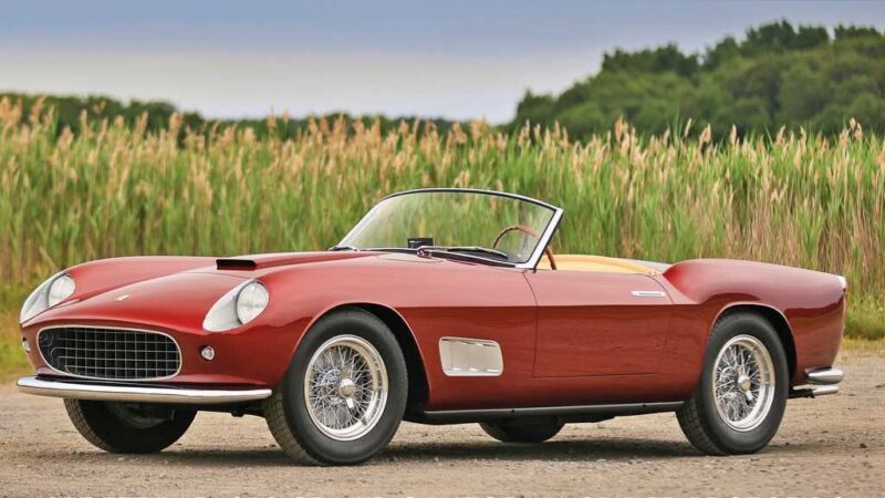 A classic red convertible sports car with chrome wire wheels, possibly featured among the 10 Most Expensive Cars To Sell At 2025 Monterey Car Week Auctions, is parked on a gravel surface in front of tall green grass and trees.