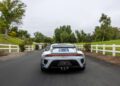 A white Porsche 935 with blue and red stripes drives down a paved road bordered by white fences and greenery on a cloudy day—a true dream car in motion.