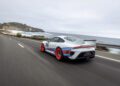 A white Porsche 935 with red wheels and racing stripes drives on a coastal highway beside the ocean under a cloudy sky—truly a dream car in motion.