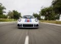 A white Porsche 935 with blue and red racing stripes drives on a road, photographed from the front at a low angle—a true dream come true for car enthusiasts. Trees and greenery create a vibrant backdrop.