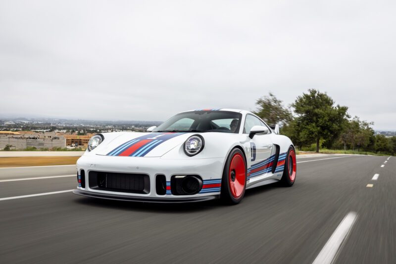 A Porsche 935 sports car with blue and red racing stripes and red wheels drives on an open road under a cloudy sky—a true dream car in motion.