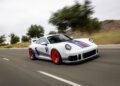 A white Rezvani Retro RR1 sports car with blue and red racing stripes and red wheels drives on a road past trees and hills on a cloudy day—truly a dream car in motion.