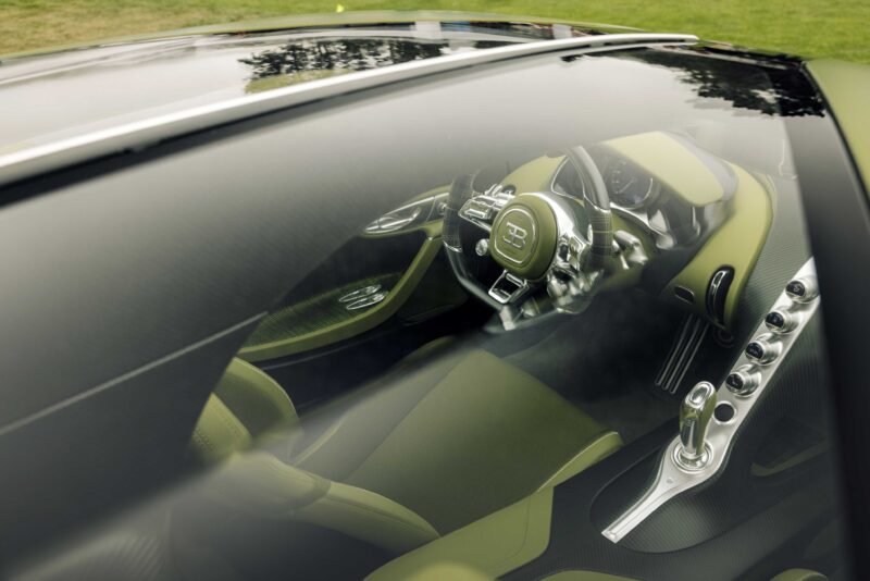 View through the windshield of a luxury car at The Quail, showcasing the green and black interior, W16 steering wheel with Bugatti Brouillard logo, and a modern central console with multiple controls.