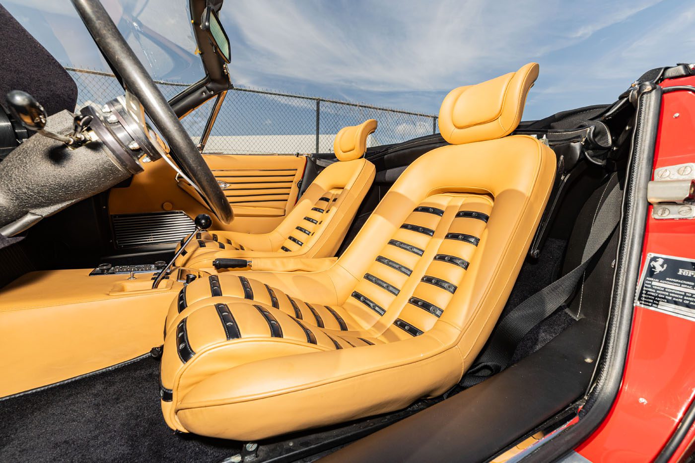 Close-up view of the tan leather interior and bucket seats of a classic red convertible sports car—one of the Ferrari Legends set to shine at Mecum Auction, Monterey 2025. Blue sky and clouds fill the backdrop.