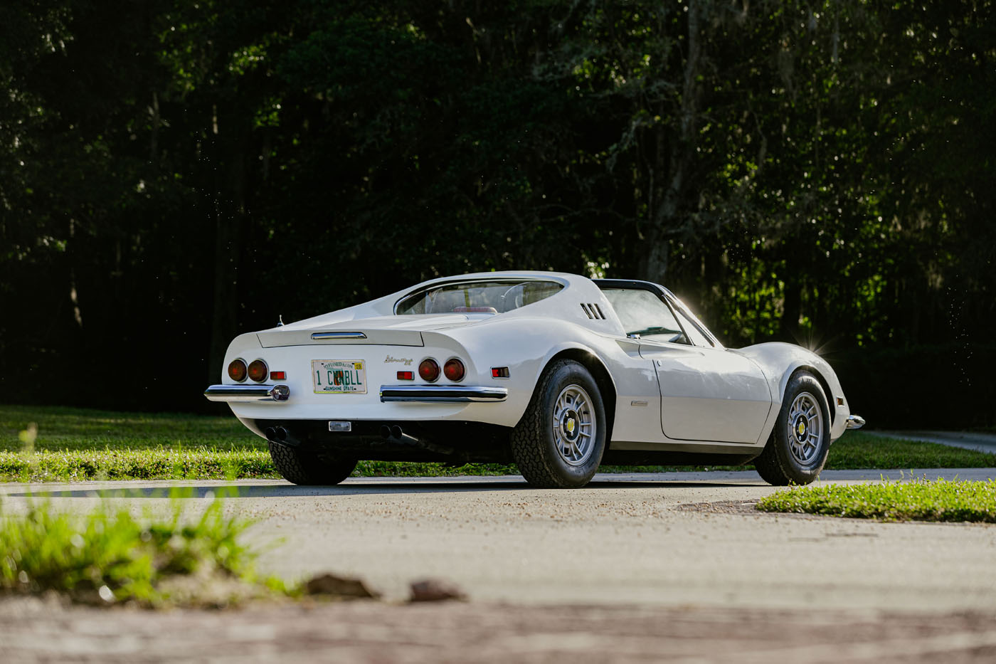 A white classic sports car—perfect for Ferrari Legends—rests on a paved driveway with trees and greenery in the background, viewed from the rear left side.