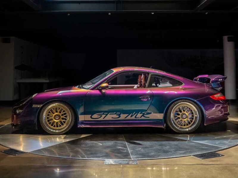 A purple and teal 997 GT3 MR sports car with gold wheels is parked indoors under spotlights, evoking a Grello Tribute at Werks Reunion Monterey.