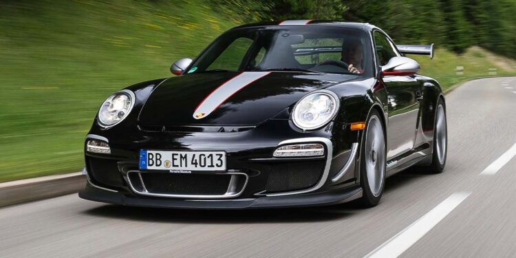 A black 997 Porsche 911 GT3 RS 4.0 with red and white stripes drives on a winding road bordered by greenery and trees.