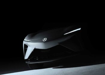 A dark, shadowed view of the front end of an Acura RSX Prototype EV Set for Global Debut at Monterey, featuring illuminated slim headlights and the iconic Acura logo.
