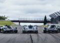 Three black hypercars, including an Aston Martin Valkyrie, are parked side by side at Monticello Raceway, with a bridge and safety barriers in the background under a cloudy sky.
