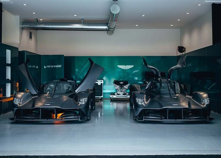 Two matte black Aston Martin Valkyries with their doors open are parked side by side in a modern garage, possibly at Monticello Raceway, with an engine displayed between them in the background.