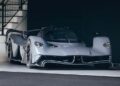 A sleek silver Aston Martin Valkyrie hypercar is parked indoors under a staircase, featuring aerodynamic design and futuristic details.