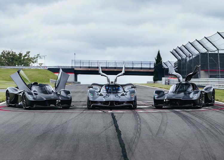 Three high-performance sports cars, including an Aston Martin Valkyrie, are parked side by side with their doors open at Monticello Raceway, with a bridge and fencing visible in the background.