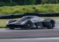 A matte black Aston Martin Valkyrie with aerodynamic design is driving on Monticello Raceway, surrounded by greenery and a barrier in the background.