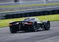 An Aston Martin Valkyrie hypercar speeds along Monticello Raceway, its tail lights glowing as it races past green grass, captured from the rear.