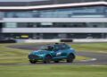A blue Aston Martin SUV with "F1" markings and a roof light bar drives on a racetrack with a modern building in the background.