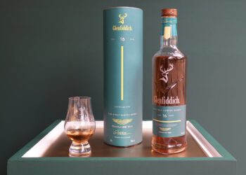 A bottle of Glenfiddich 16-year-old single malt Scotch whisky, inspired by Aston Martin’s new Formula 1 Team Scotch Whisky, with its cylindrical box and a glass of whisky on a white tray against a green background.