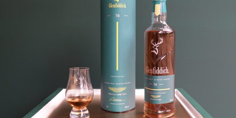 A bottle of Glenfiddich 16-year-old single malt Scotch whisky, inspired by Aston Martin’s new Formula 1 Team Scotch Whisky, with its cylindrical box and a glass of whisky on a white tray against a green background.