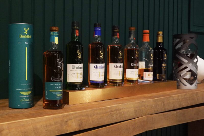 A row of Glenfiddich whisky bottles, including the Tasted: Aston Martin’s New Formula 1 Team Scotch Whisky by Glenfiddich, are displayed on a wooden surface against a dark green wall.