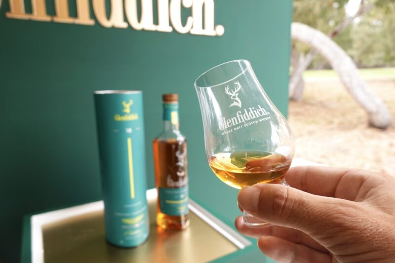 A hand holds a Glenfiddich whisky glass with amber liquid; in the background, a Glenfiddich bottle and box are displayed, celebrating Tasted: Aston Martin’s New Formula 1 Team Scotch Whisky by Glenfiddich.