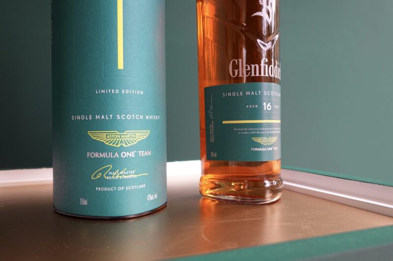 A bottle and cylindrical box of Glenfiddich 16 Year Old Single Malt Scotch Whisky, Tasted: Aston Martin’s New Formula 1 Team Scotch Whisky by Glenfiddich, are displayed on a reflective surface against a green background.