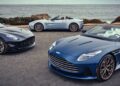 Three blue Aston Martin sports cars, including two convertibles and a coupe, are parked near a coastal road with the ocean beyond—celebrating Aston Martin Marks 75 Years in the Americas With Thrillseeker Collection and Valhalla Debut at Monterey.