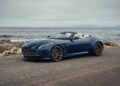 A blue convertible sports car, reminiscent of the Aston Martin Marks 75 Years in the Americas, is parked on a coastal road overlooking the ocean and rocky shoreline beneath a cloudy sky.