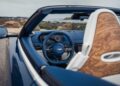 View of a luxury convertible car's interior—blue leather steering wheel, digital dashboard, and wood-trimmed seat backs—with an ocean landscape in the background, inspired by Aston Martin Marks 75 Years in the Americas With Thrillseeker Collection.