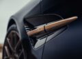 Close-up of a car's side vent with a gold accent bar labeled "V12," showcasing the smooth, dark bodywork—part of Aston Martin’s Thrillseeker Collection marking 75 years in the Americas at Monterey.