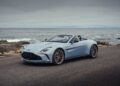 A light blue convertible sports car, inspired by Aston Martin marking 75 years in the Americas with its Thrillseeker Collection and Valhalla debut at Monterey, is parked on a coastal road with the ocean and cloudy sky in the background.