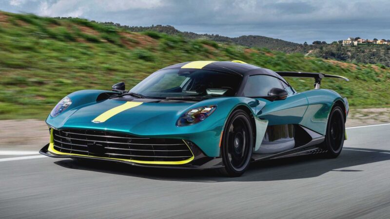 A green and yellow sports car, inspired by Aston Martin's Thrillseeker Collection marking 75 years in the Americas, speeds along a road with lush greenery and rolling hills in the background.