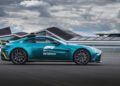 A teal Aston Martin Vantage S, serving as the official Formula 1 Safety Car with F1 and Aramco branding, is parked on a race track with modern buildings and a cloudy sky in the background.
