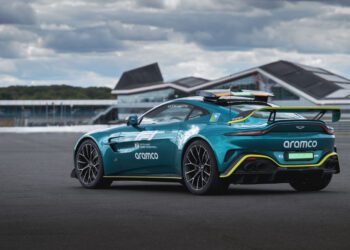 A teal Aston Martin Vantage S Formula 1 Safety Car with Aramco branding is parked on a racetrack, set against modern buildings and a cloudy sky in the background.