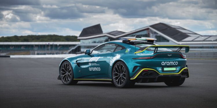 A teal Aston Martin Vantage S Formula 1 Safety Car with Aramco branding is parked on a racetrack, set against modern buildings and a cloudy sky in the background.