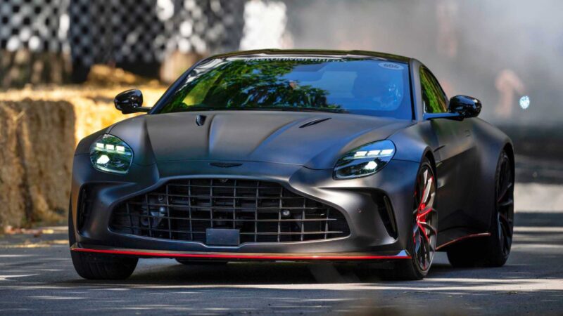 A matte black Aston Martin sports car with red accents drives on a paved track, showcasing the Thrillseeker Collection as Aston Martin marks 75 years in the Americas, surrounded by hay bales and a blurred background.