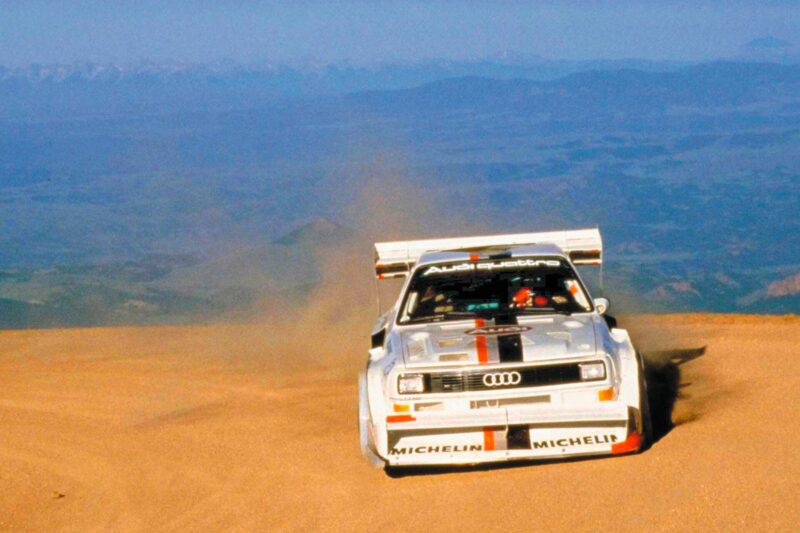An Audi Quattro rally car, powered by the last surviving 5-cylinder on sale today that packs a punch, races up a dirt road on a mountain, kicking up dust amid a scenic valley and distant mountains in the background.
