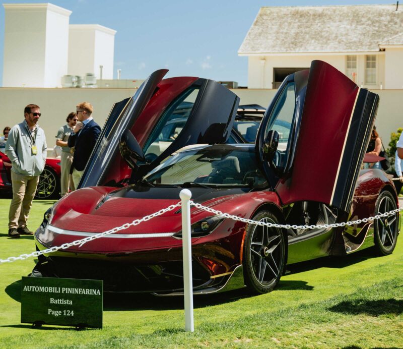 A red Automobili Pininfarina Battista with butterfly doors open is displayed outdoors on grass, surrounded by people and a white chain barrier, as Automobili Pininfarina closes the Battista chapter with the one-off Novantacinque at Pebble Beach.