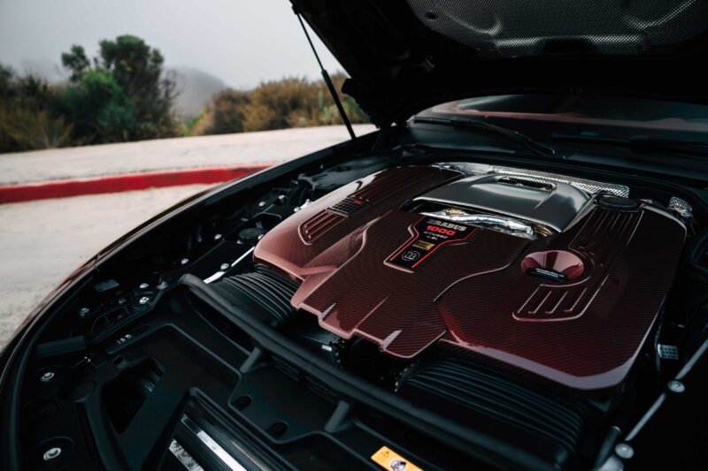 Close-up view of a high-performance V8 Biturbo engine under the hood of a car, similar to the BRABUS Rocket GTC Deep Red that breaks cover at Pebble Beach with 1000 HP and an all-carbon body, with the landscape visible in the background.