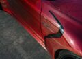 Close-up view of the BRABUS Rocket GTC Deep Red breaks cover at Pebble Beach, highlighting its carbon fiber texture, black trim, and "V8 BITURBO" badge on an all-carbon body parked on dark asphalt.