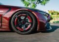 Close-up of the front side of a shiny red BRABUS Rocket GTC Deep Red, featuring a large black alloy wheel and carbon fiber detailing, parked on a sunlit street. This striking car breaks cover at Pebble Beach with 1000 HP and an all-carbon body.