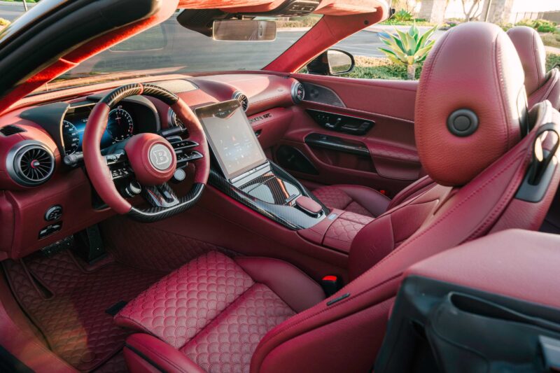 Luxury car interior featuring red leather seats, a detailed dashboard with digital displays, and a large central touchscreen. Inspired by the BRABUS Rocket GTC Deep Red Breaks Cover at Pebble Beach, this convertible exudes power and sophistication.