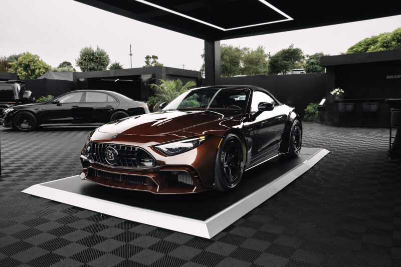 A red BRABUS-modified Mercedes-Benz convertible is displayed on a platform in a modern showroom, evoking the elegance of Pebble Beach, with another black car in the background.