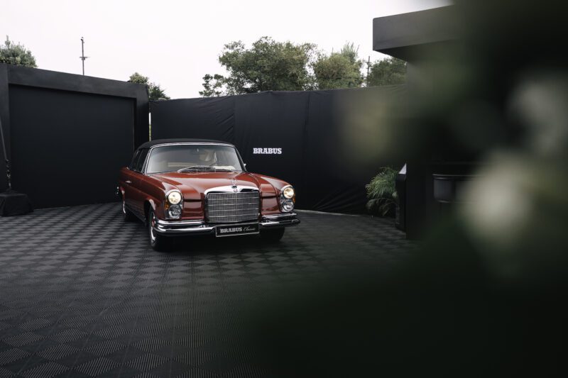 A red vintage Mercedes-Benz car is parked on a checkered black floor in front of a BRABUS display area at Pebble Beach, with lush greenery in the background.